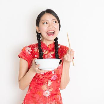 Portrait of young Asian woman in traditional cheongsam dress eating, hand holding bowl and chopsticks, celebrating Chinese Lunar New Year or spring festival, standing on plain background.