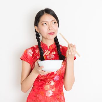 Portrait of young Asian girl in traditional cheongsam dress eating, hand holding bowl and chopsticks, celebrating Chinese Lunar New Year or spring festival, standing on plain background.