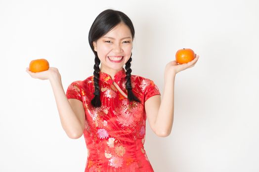 Young Asian woman in traditional qipao dress holding mandarin orange and smiling, celebrating Chinese Lunar New Year or spring festival, standing on plain background.