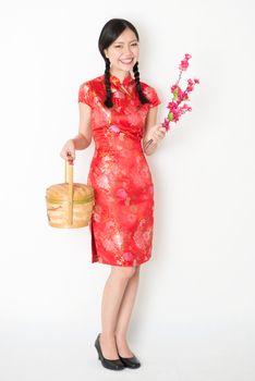 Young Asian woman in traditional qipao dress holding gift basket and plum blossom smiling, celebrating Chinese New Year or spring festival, full length standing on plain background.