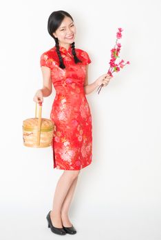 Young Asian woman in traditional cheongsam dress holding gift basket and plum blossom smiling, celebrating Chinese Lunar New Year or spring festival, full body standing on plain background.