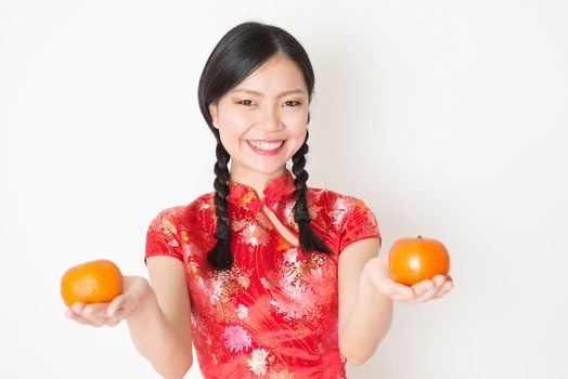 Young Asian woman in traditional cheongsam dress holding mandarin orange and smiling, celebrating Chinese Lunar New Year or spring festival, standing on plain background.