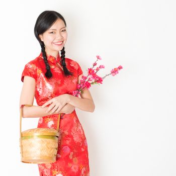 Young Asian woman in traditional cheongsam dress holding gift basket and plum blossom smiling, celebrating Chinese Lunar New Year or spring festival, standing on plain background.