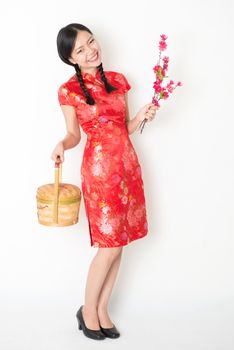Young Asian woman in traditional cheongsam dress holding gift basket and plum blossom smiling, celebrating Chinese Lunar New Year or spring festival, full length standing on plain background.