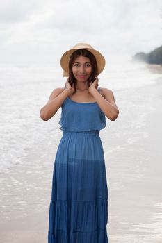 portrait of smiling face of young asian tan skin woman with long dress and fashion wide straw hat standing on sea beach 