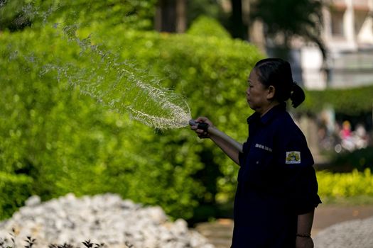 Unidentified Vietnamese woman with conical hat watering grass in a park.