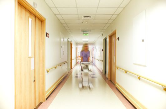 A blurred figure of nurse, coming down a well lit hospital corridor, pushing equipment.