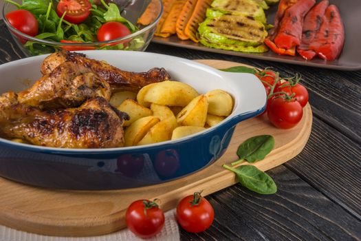 Tasty and nutritious meal with baked chicken drumsticks and potato in ceramic dish,  grilled vegetables, salad and tomatoes on the background of dark table