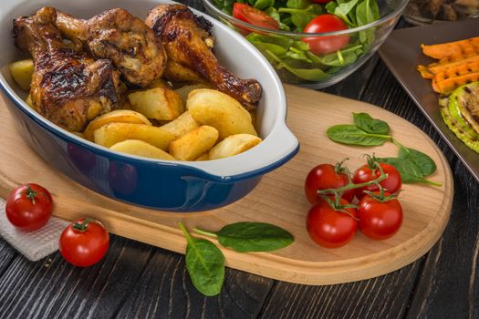 Tasty and nutritious meal with baked chicken drumsticks and potato in ceramic dish,  grilled vegetables, salad and tomatoes on the background of dark table