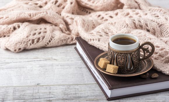 Cup of coffee on a notebook with beige knitted scarf and sugar lying on a white table
