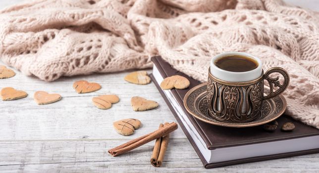 Cup of coffee on a notebook with beige knitted scarf ,cinnamon and a lot of wooden hearts lying on a white table