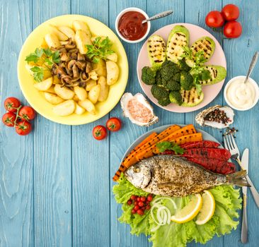 Delicious and nutritious meal rich in vitamins and minerals with fish and grilled vegetables: potatoes, mushrooms, carrot, tomato, broccoli, pepper, squash,paprika, lemon on a blue wooden table.