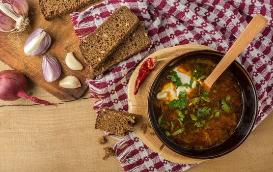 Tasty and nutritious soup with meat, potatoes, cabbage, tomatoes, beans, sour cream, parsley, onion, garlic, dark bread in a clay plate on a wooden tray and wooden table. Top view