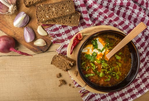 Tasty and nutritious soup with meat, potatoes, cabbage, tomatoes, beans, sour cream, parsley, onion, garlic, dark bread in a clay plate on a wooden tray and wooden table. Top view