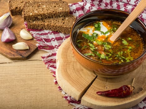 Tasty and nutritious soup with meat, potatoes, cabbage, tomatoes, beans, sour cream, parsley, onion, garlic, dark bread in a clay plate on a wooden tray and wooden table.