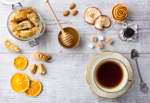 Tasty and healthy almond cookies, rich in vitamins, minerals in a glass jar, cup of tea, honey, almonds, chocolates, dried apples, dried lemons and sugar on white wooden table. Top view.