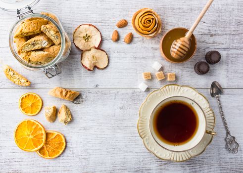 Tasty and healthy almond cookies, rich in vitamins, minerals in a glass jar, cup of tea, honey, almonds, chocolates, dried apples, dried lemons and sugar on white wooden table. Top view.