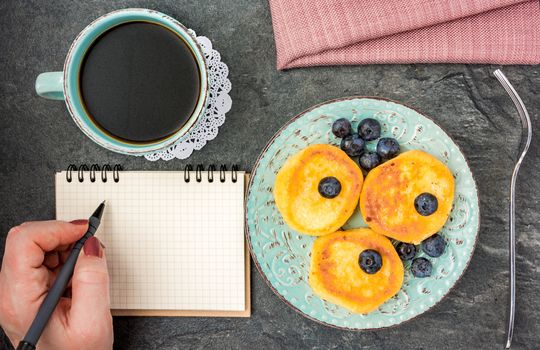 Woman having breakfast with cheese pancake with blueberries, powdered sugar, black coffee on a dark wooden table and writing in a notebook . Top view.