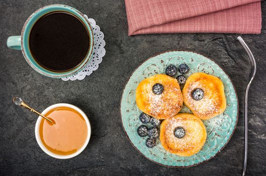 cheese pancake with blueberries, powdered sugar, honey and black coffee on a dark background top view