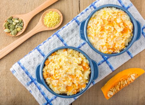 millet porridge with pumpkin in blue bowl on a kitchen towel, pumpkin seeds and millet in a wooden spoons and pumpkin on a wooden table top view