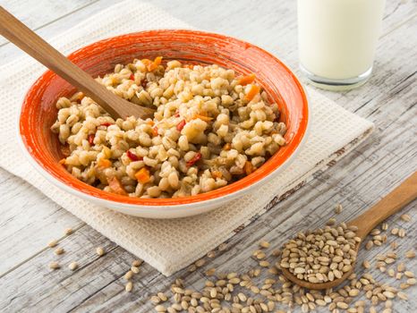 Pearl barley porridge with vegetables in an orange ceramic plate with a kitchen towel, pearl barley in a wooden spoon and glass of milk on a white wooden table