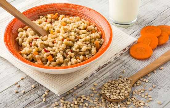 Pearl barley porridge with vegetables in an orange ceramic plate with a kitchen towel, pearl barley in a wooden spoon and glass of milk on a white wooden table