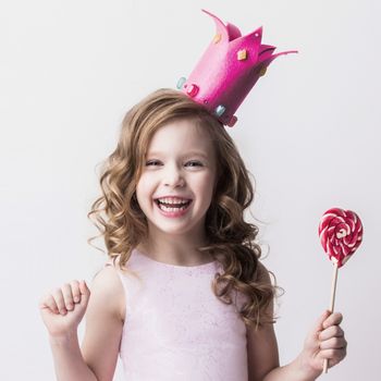 Beautiful little candy princess girl in crown holding big pink heart lollipop and smiling