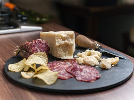 A dark stone tray on a wooden kitchen table filled with parmesan cheese, salami and potato chips.