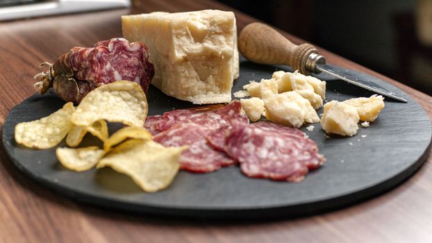 A dark stone tray on a wooden kitchen table filled with parmesan cheese, salami and potato chips.