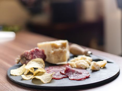 A dark stone tray on a wooden kitchen table filled with parmesan cheese, salami and potato chips.