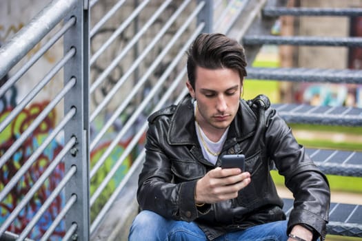 Handsome young man standing outdoors in urban environment on metal stairs, using cell phone