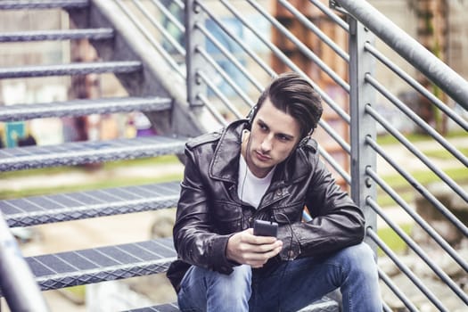 Handsome young man standing outdoors in urban environment on metal stairs, using cell phone