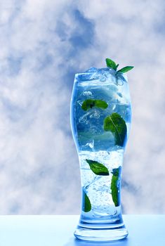 Minty refreshing drink in glass with ice and leaves on a background of blue summer sky and clouds