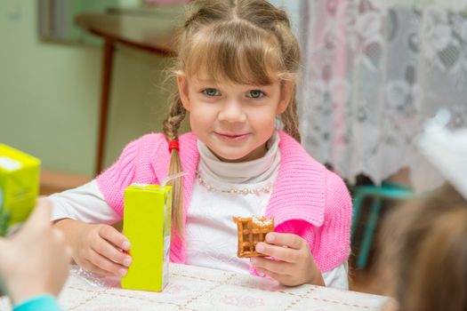 The girl is drinking juice and eating cookies at the desk in the kindergarten