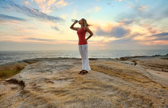 Female drinking bottled water outdoors by the ocean with pretty sky behind her