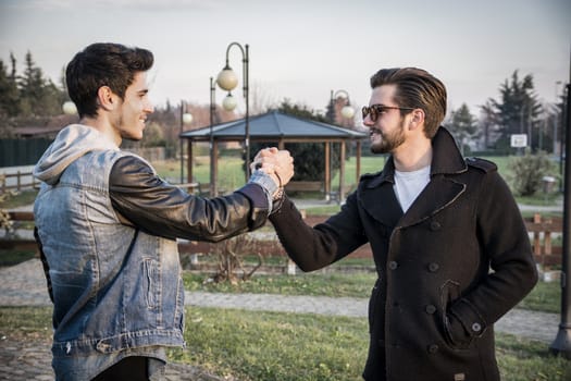 Two handsome casual trendy young men greeting outdoors in an urban park gripping hands with happy welcoming smiles