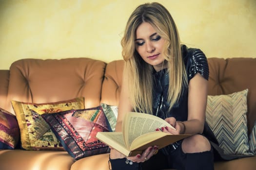 Portrait of a beautiful blond woman in black dress posing for camera at home, sitting and reading a book