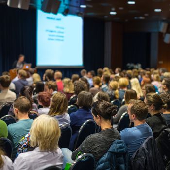 Business and entrepreneurship symposium. Speaker giving a talk at business meeting. Audience in the conference hall. Rear view of unrecognized participant in audience.