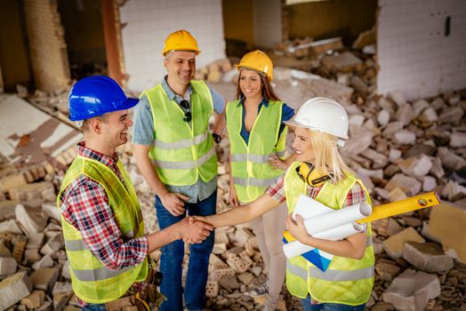 Young female construction architects and electrician shaking hands in the building damaged in the disaster while in the background standing theirs colleagues.