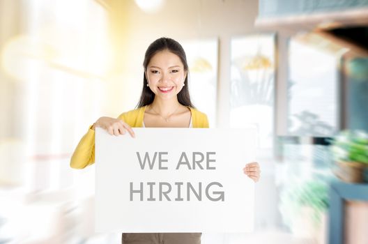 Young Asian woman hand holding "we are hiring" board sign in restaurant or cafe. 