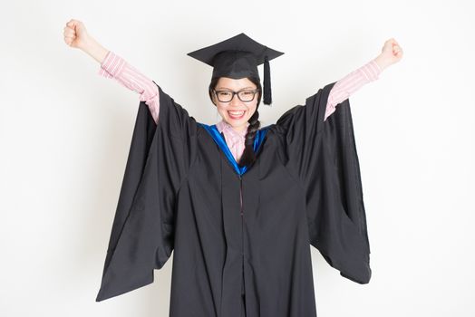 Happy university student in graduation gown and cap arms raised and smiling. Portrait of east  Asian female model standing on plain background.