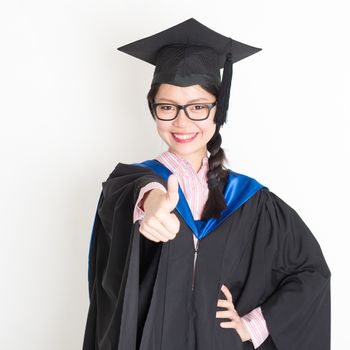 Happy university student in graduation gown and cap giving thumb up sign. Portrait of east  Asian female model standing on plain background.