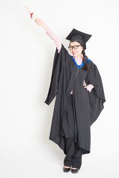 University student in graduation gown and cap hand raised holding diploma certificate. Full body portrait of east  Asian female model standing on plain background.