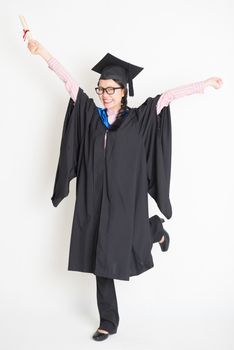 University student in graduation gown and cap hand raised holding diploma certificate jumping around. Full body portrait of east  Asian female model standing on plain background.