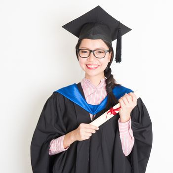 Happy university student in graduation gown and cap holding diploma certificate. Portrait of east  Asian female model standing on plain background.