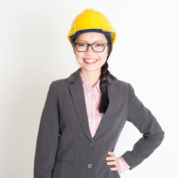 Portrait of Asian female engineer with hard hat smiling and looking at camera, standing on plain background.