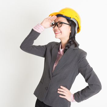 Portrait of Asian female engineer with hard hat hand shielding and looking away, standing on plain background.