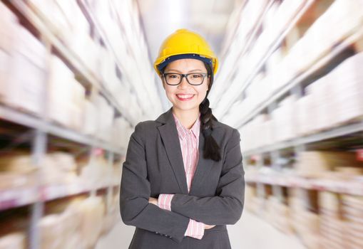 Young Asian female store manager with hard hat arms crossed standing in storehouse. Shelves with goods at background.