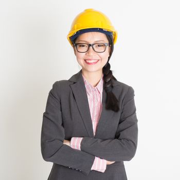 Portrait of Asian female architect with hard hat smiling and looking at camera, standing on plain background.