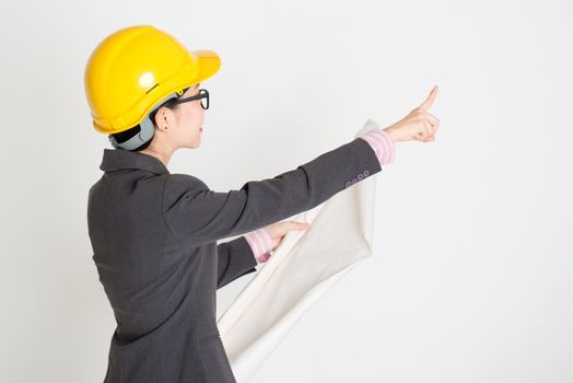 Portrait of Asian female contractor engineer with hard hat holding blue print and pointing away, standing on plain background.